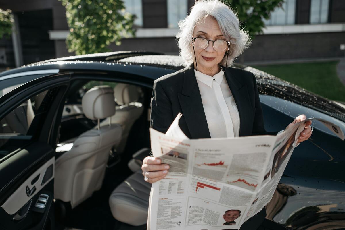 A woman reading a newspaper beside her car